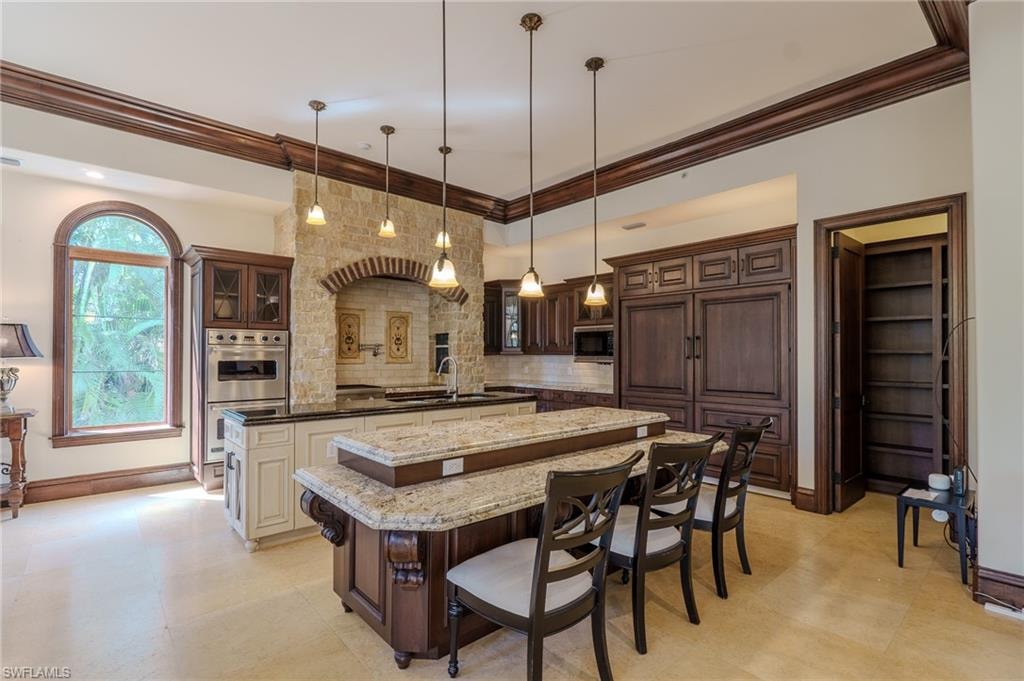 Dual tone kitchen with a kitchen breakfast bar, dark stone counters, double oven, glass insert cabinets, and hanging light fixtures