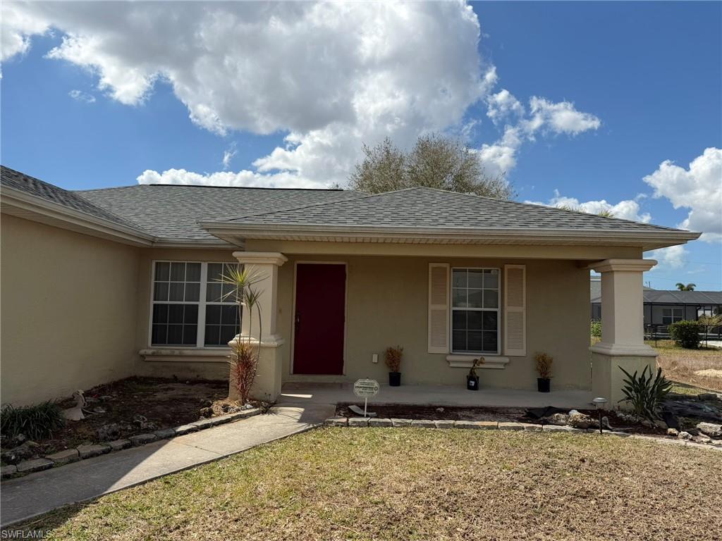 View of exterior entry featuring roof with shingles, stucco siding, a porch, and a lawn