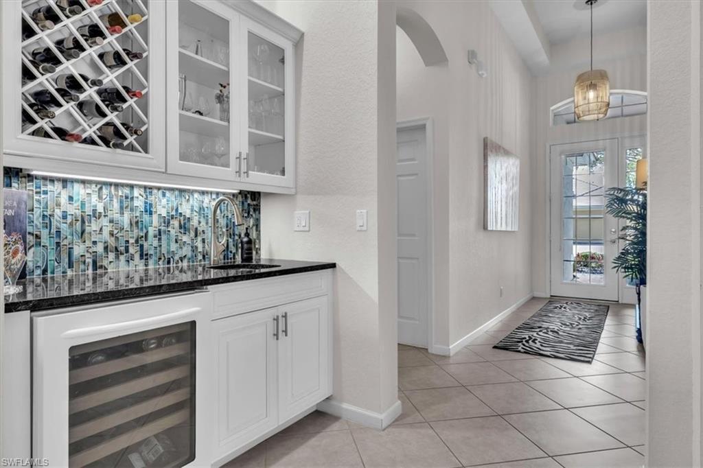 Indoor wet bar featuring white cabinetry, beverage cooler, dark stone countertops, glass insert cabinets, and arched walkways