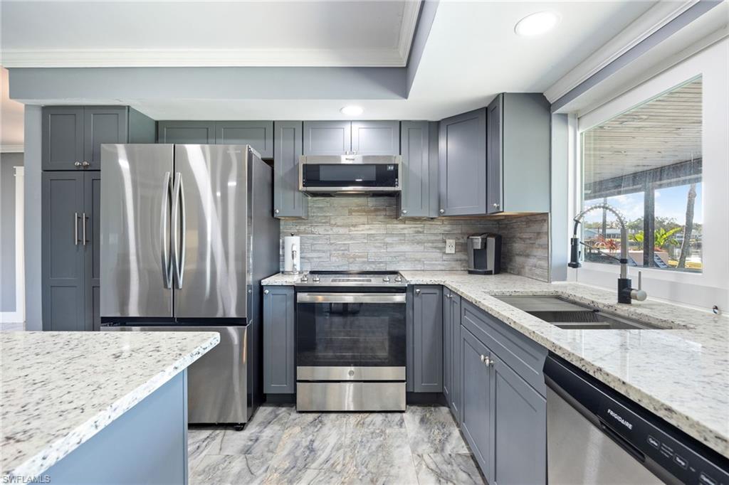 Kitchen featuring stainless steel appliances, light stone countertops, crown molding, gray cabinetry, and decorative backsplash