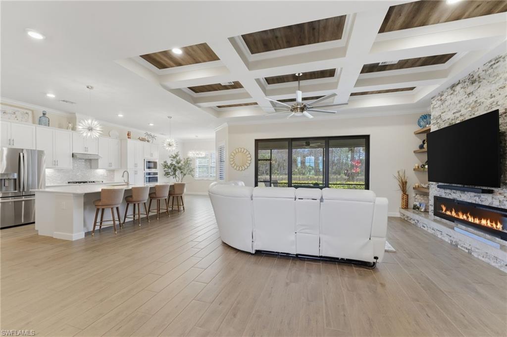 Living room with ceiling fan, a stone fireplace, coffered ceiling, light wood finished floors, and suspended lighting