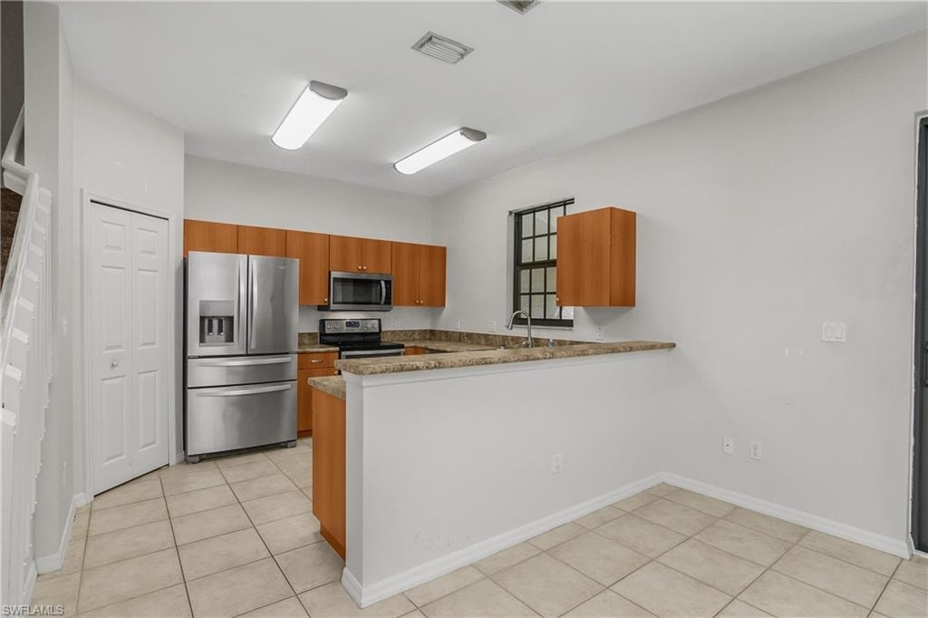 Kitchen featuring stainless steel appliances, wood finish cabinets, a peninsula, light tile patterned floors, and dark stone counters