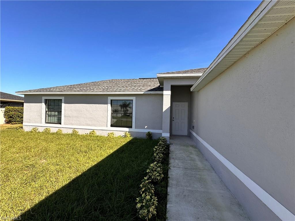 View of exterior entry with a shingled roof, a lawn, and stucco siding