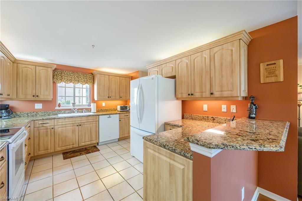 Kitchen with light wood finish cabinets, light stone countertops, a peninsula, white appliances, and a kitchen bar