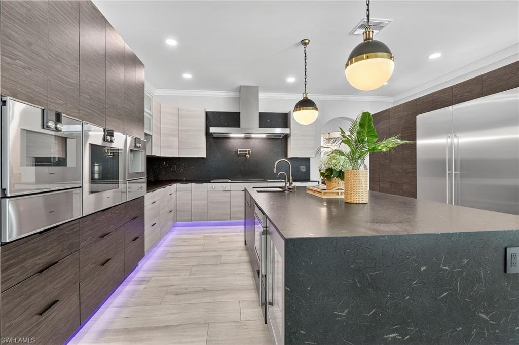 Kitchen with a large island with sink, stainless steel appliances, a warming drawer, decorative light fixtures, and dark stone counters
