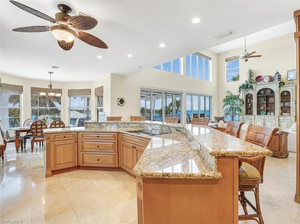 Kitchen with a ceiling fan, a breakfast bar area, light stone countertops, a spacious island, and a high ceiling - Virtually Edited Image