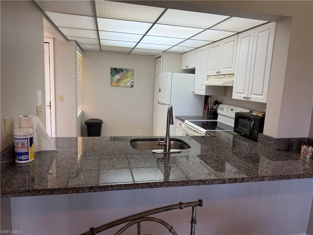 Kitchen featuring a peninsula, white cabinetry, white appliances, a paneled ceiling, and dark stone countertops