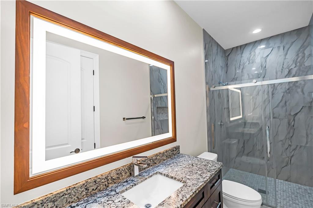 Bathroom featuring an illuminated wood-framed mirror, a stone-finish countertop with an undermount sink, a dark wood-finish vanity, a glass-enclosed shower with textured wall tile, and a patterned shower floor