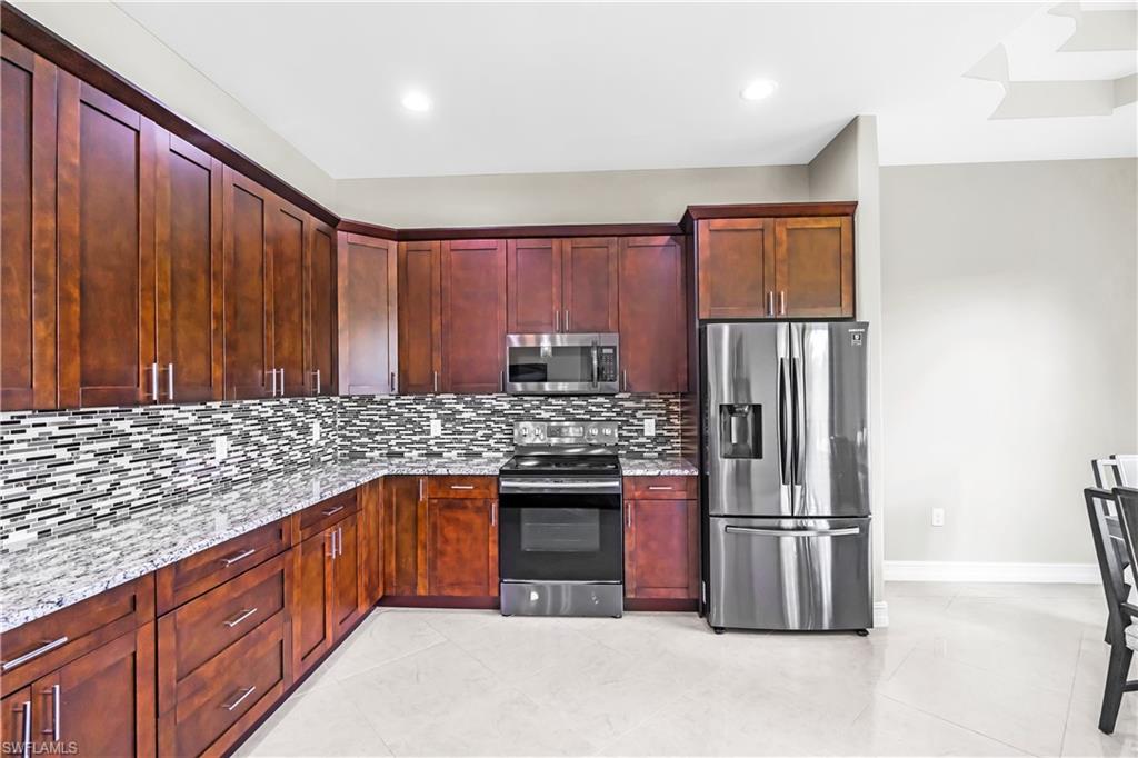 Open-concept kitchen featuring dark wood cabinetry, stainless steel appliances, speckled stone countertops, a mosaic tile backsplash, and light tile flooring