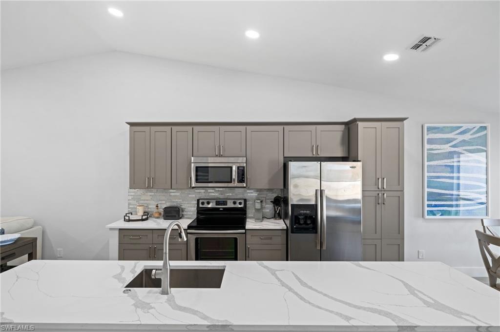 Kitchen featuring gray cabinets, stainless steel appliances, light stone countertops, vaulted ceiling, and decorative backsplash
