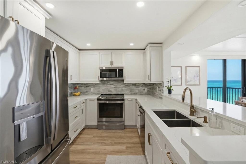 Modern kitchen featuring stainless steel appliances, white cabinetry with bronze hardware, light-toned countertops, and wood-finish flooring - Virtually Edited Image