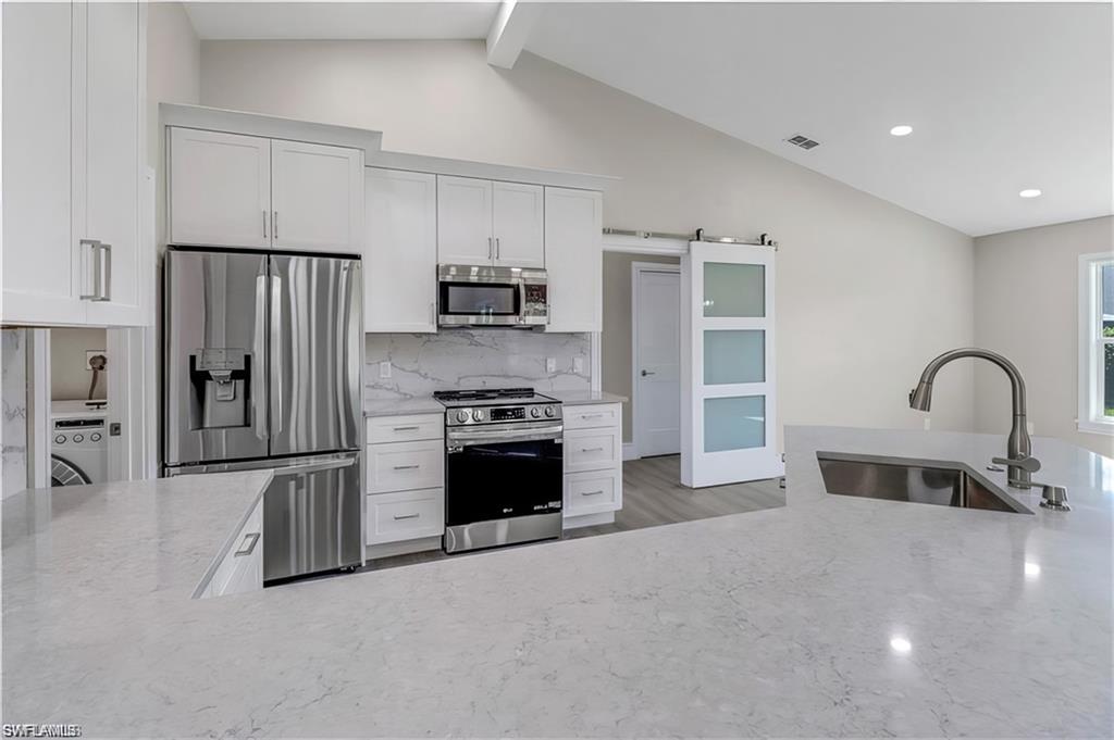 Kitchen with stainless steel appliances, a barn door, white cabinetry, light stone counters, and vaulted ceiling with beams