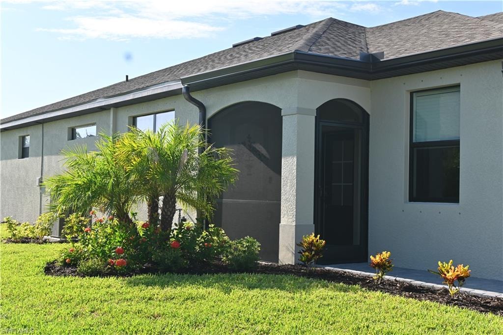 View of exterior entry with a shingled roof, stucco siding, and a lawn