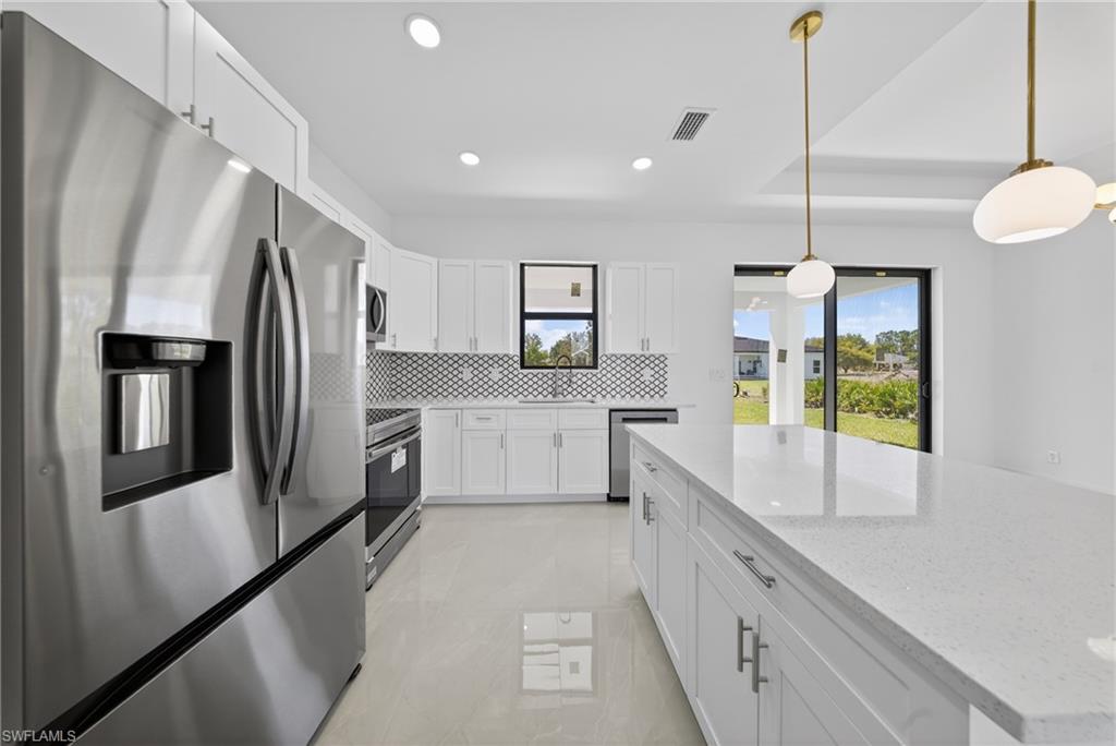 Kitchen with stainless steel appliances, white cabinets, light stone counters, backsplash, and decorative light fixtures