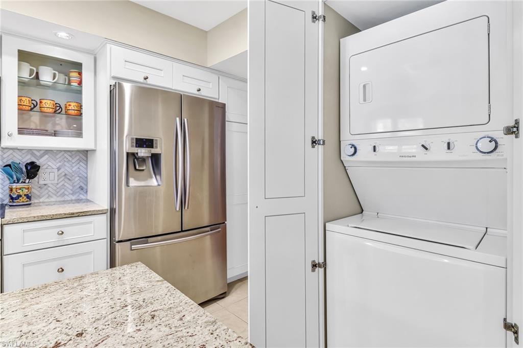 Kitchen featuring stainless steel fridge with ice dispenser, white cabinets, light stone counters, stacked washer / dryer, and glass insert cabinets