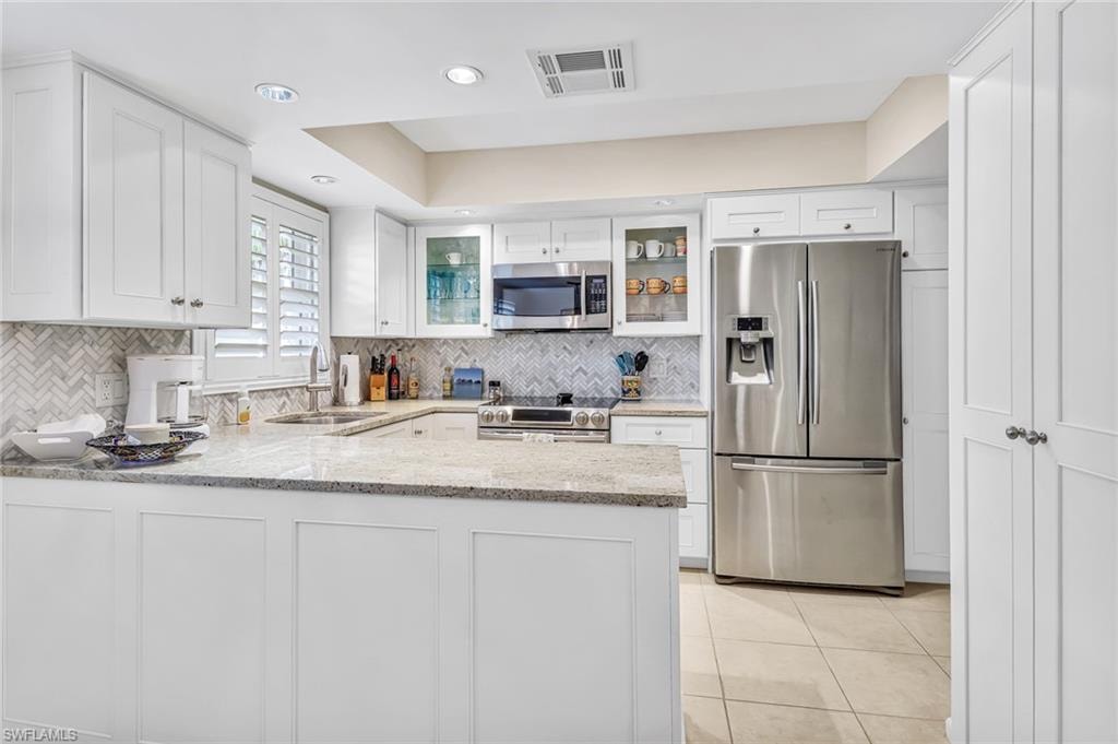 Kitchen with stainless steel appliances, a peninsula, light stone counters, glass fronted cabinets, and white cabinets