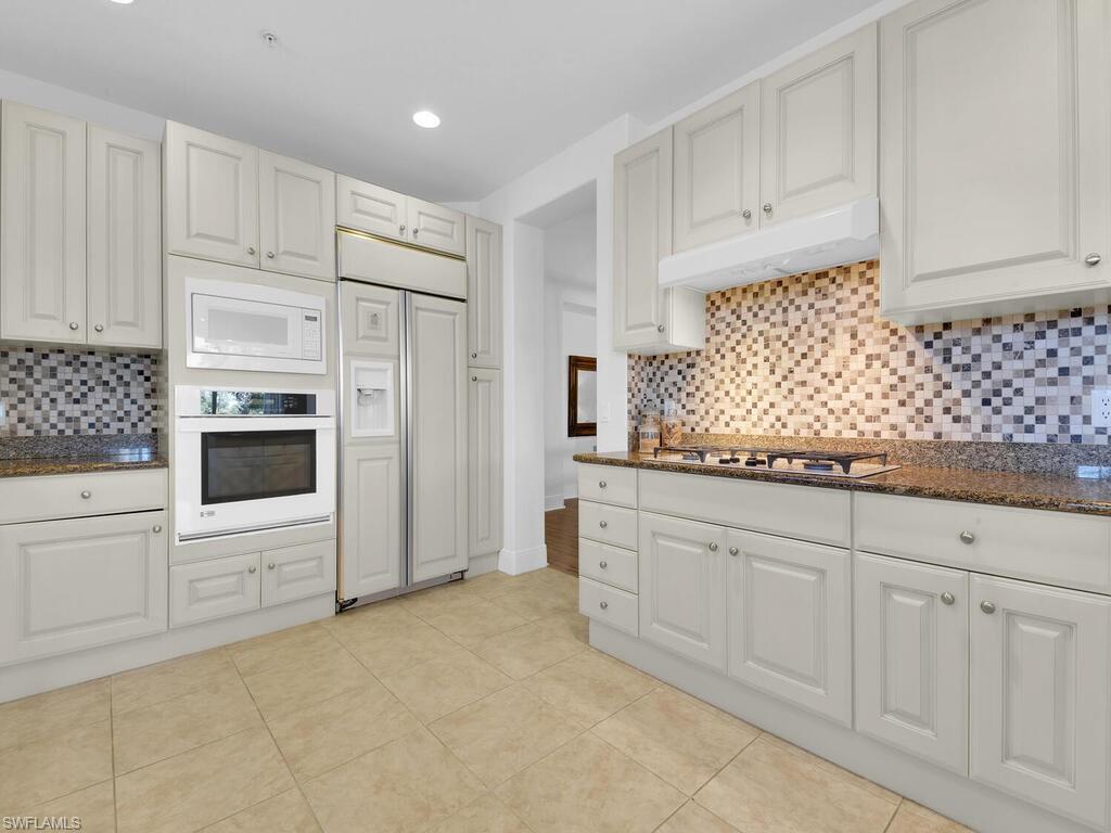 Kitchen featuring built in appliances, dark stone countertops, under cabinet range hood, and backsplash