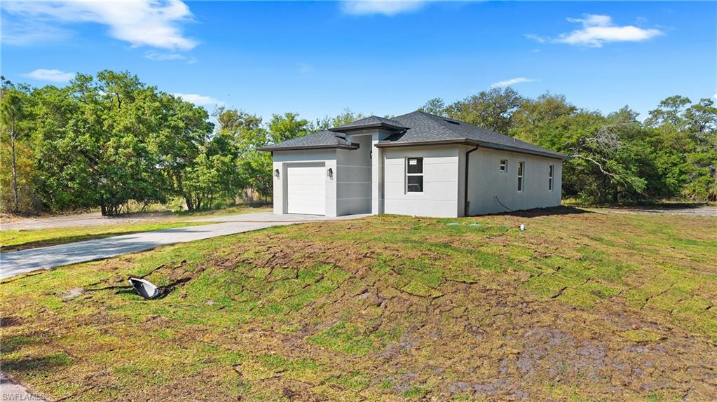 View of property exterior with an attached garage, concrete driveway, a lawn, stucco siding, and view of wooded area