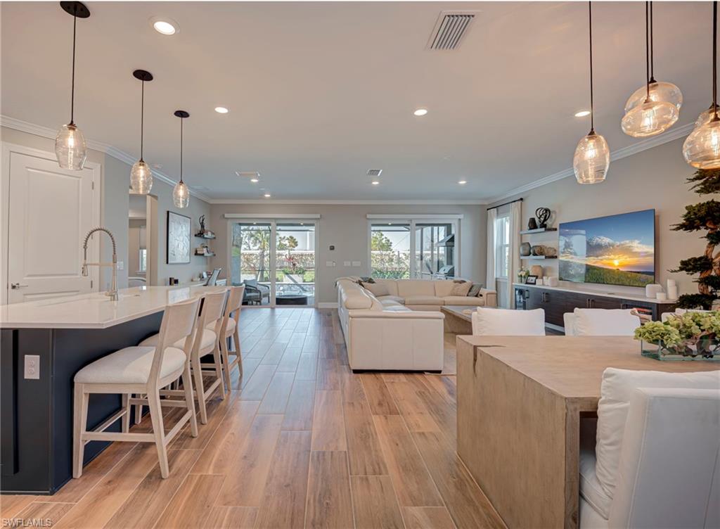 Light-filled open floor plan showcasing a designer kitchen with a large island, stylist pendant lights, and ample seating overlooking the expansive living and dining area with beautiful flooring