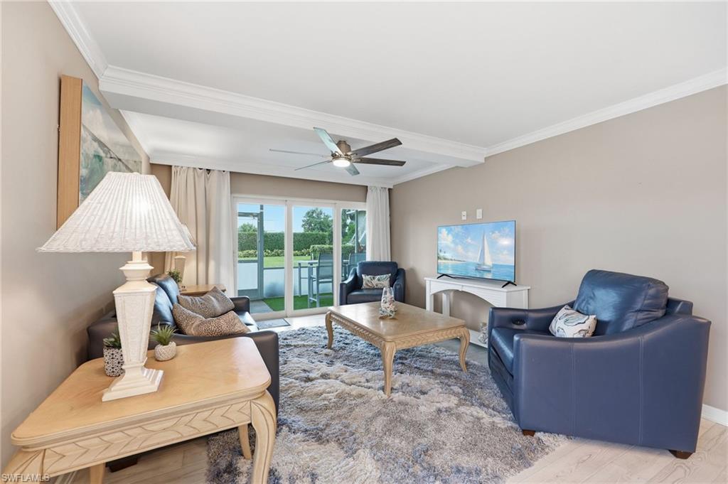 Spacious living room featuring floor-to-ceiling sliding glass doors with direct access to a covered balcony, ceiling fan, neutral wall finishes, wood-finish coffee table, plush seating, area rug, table lamp with pleated shade, and a wall-mounted flat-screen television