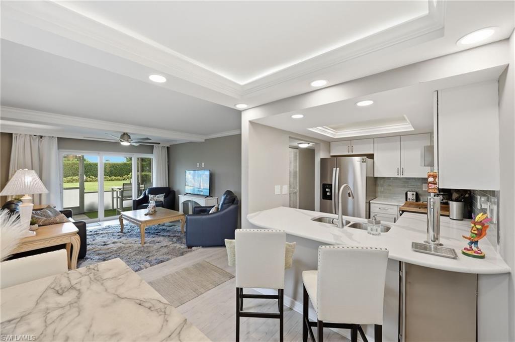 Open-concept living area featuring tray ceilings with recessed lighting, sliding glass doors to outdoor space, a kitchen island with seating, stainless steel appliances, and a tile backsplash