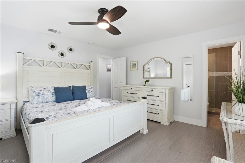Bedroom featuring light gray walls, wood-finish flooring, a ceiling fan with integrated lighting, and a doorway opening to a shower with tiled walls and a glass enclosure - Virtually Edited Image