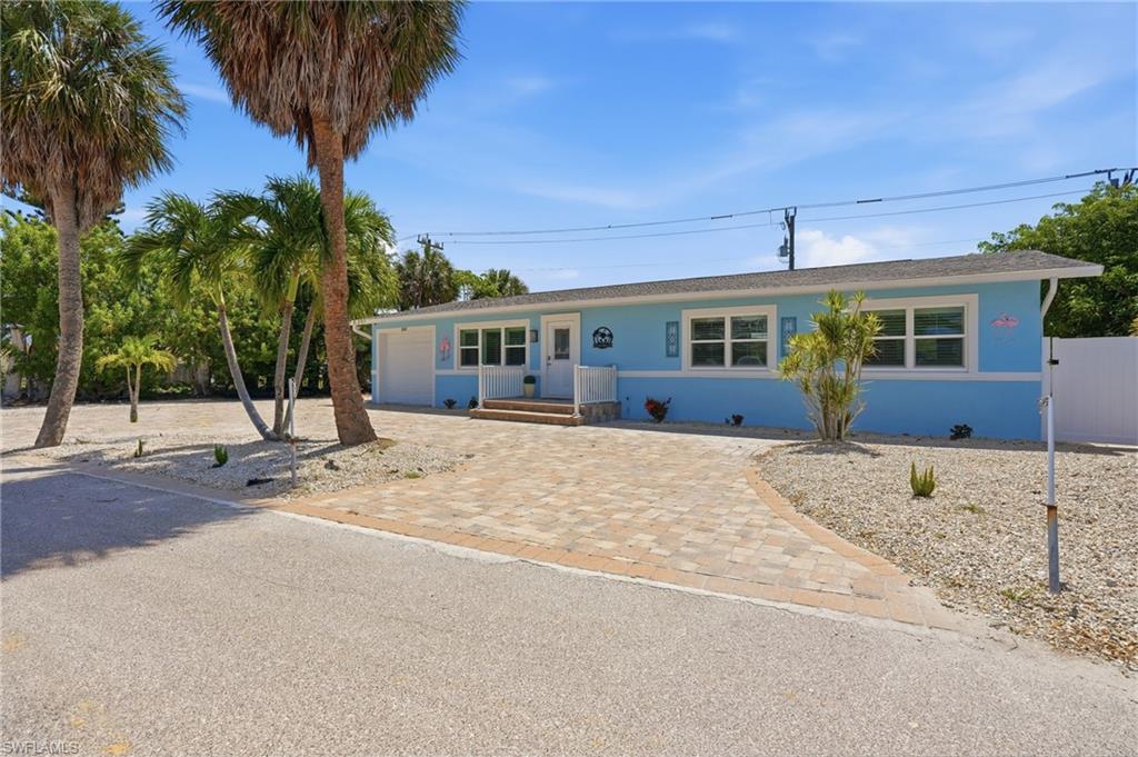 Light blue exterior with white trim, featuring a paver driveway, white privacy fence, and mature palm trees - Virtually Edited Image