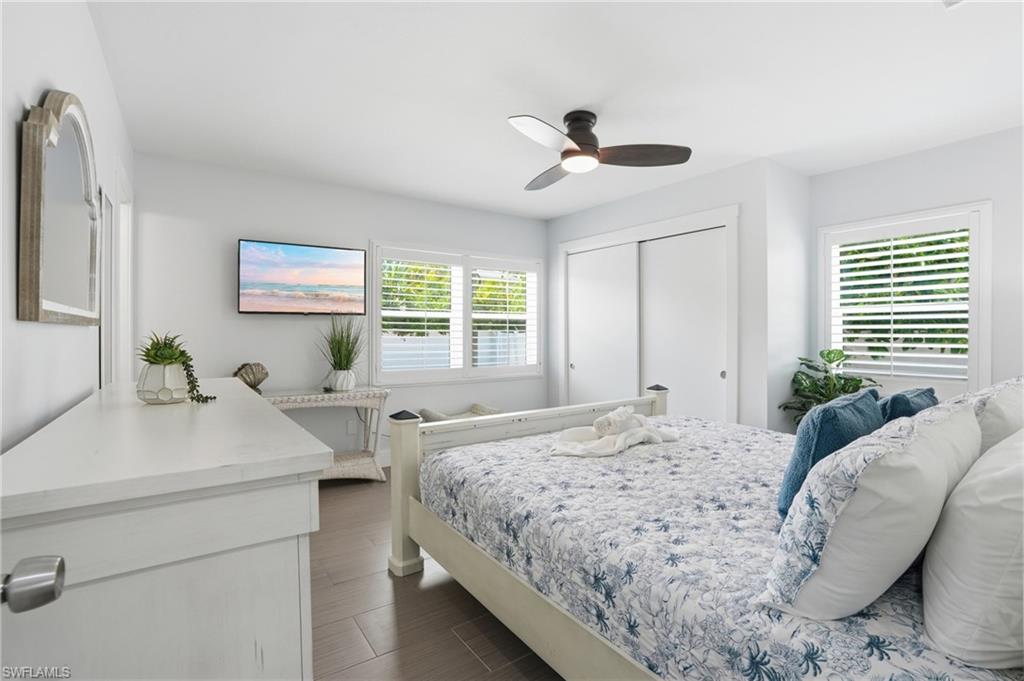 Interior room featuring wood-finish flooring, two windows with plantation shutters, a ceiling fan with integrated lighting, and a closet with sliding doors - Virtually Edited Image