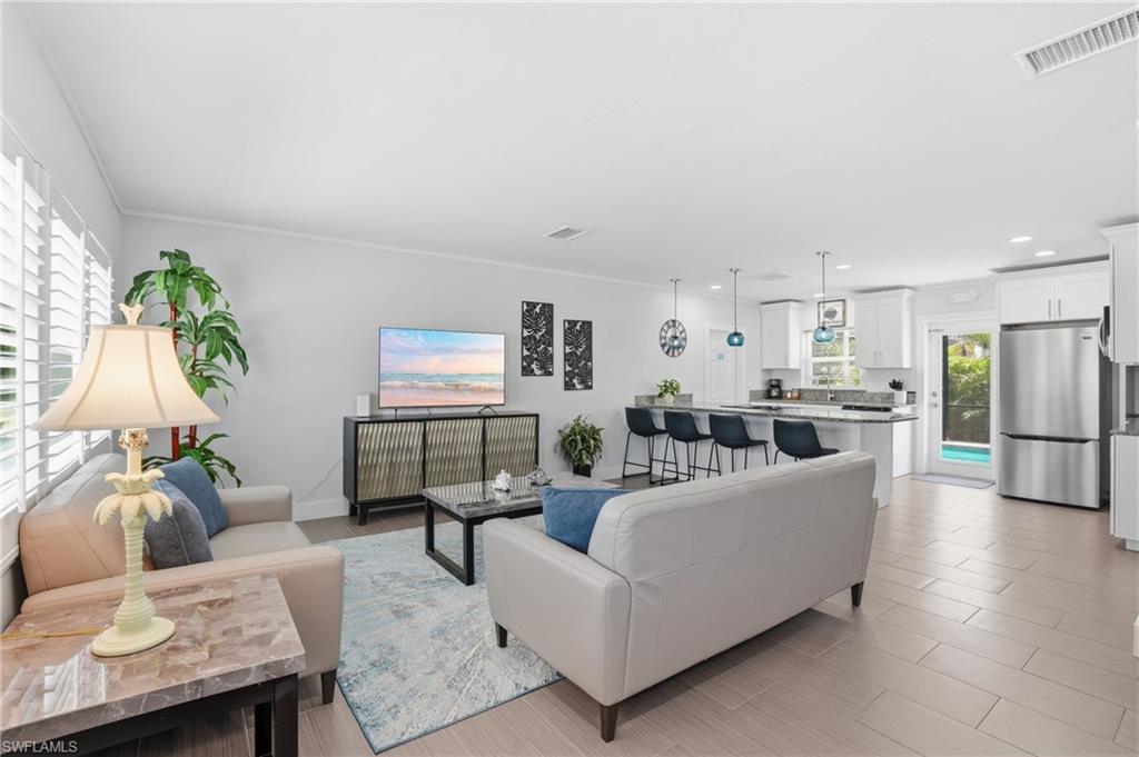 Open-concept living area featuring a kitchen with white cabinetry and stainless steel appliances, a breakfast bar with pendant lighting, and light-toned tile flooring - Virtually Edited Image