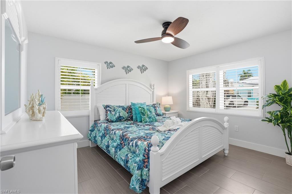 Bright bedroom featuring two windows with white louvered shutters, a white paneled bed frame, a coordinating white dresser with a mirror, a ceiling fan with integrated lighting, and wood-finish flooring - Virtually Edited Image