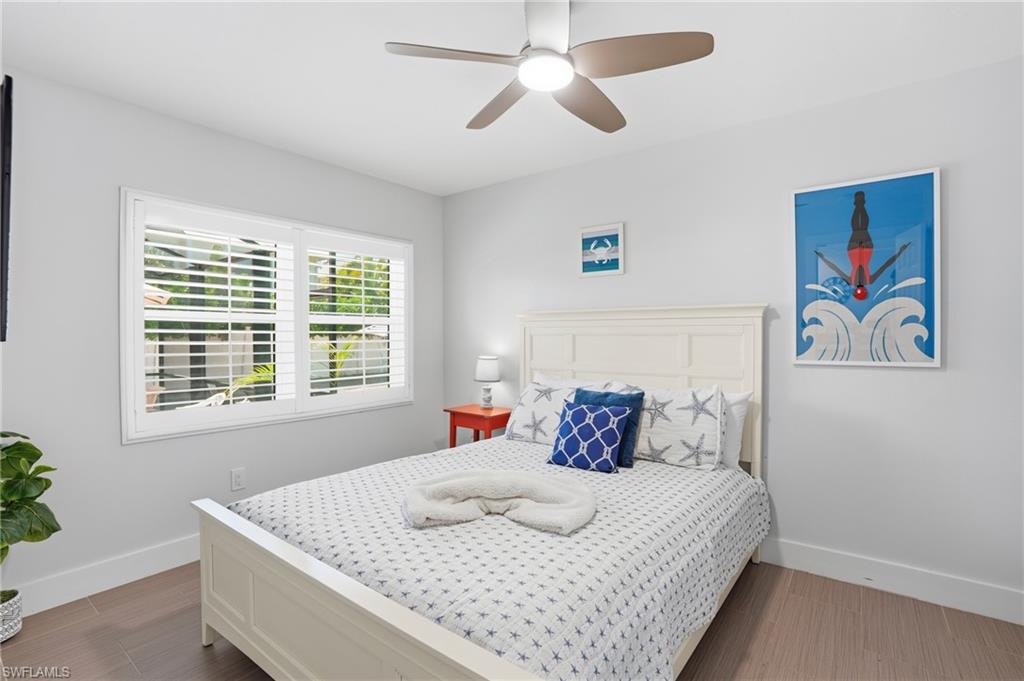 Bedroom featuring light wood-finish flooring, white walls, and a window with plantation shutters - Virtually Edited Image