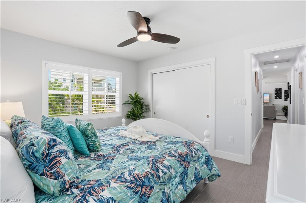 Bedroom featuring a ceiling fan with integrated lighting, a window with louvered shutters, light gray wall paint, and wood-finish flooring - Virtually Edited Image