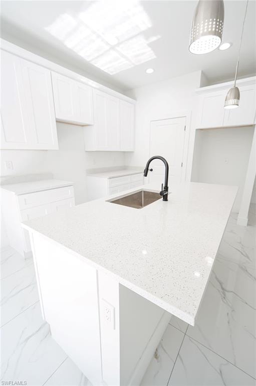 Kitchen featuring white cabinets, light stone countertops, an island with sink, hanging light fixtures, and light marble finish floors. Photos are of a staged model or similar plan. Features and finishes may vary.