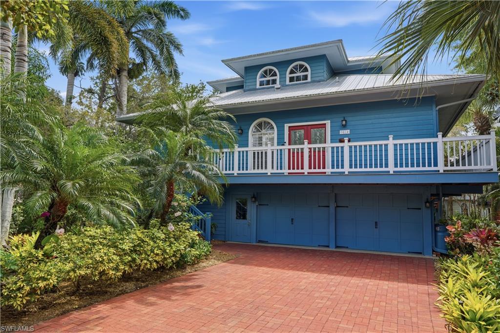 Coastal-style home featuring a blue facade with red entry doors, a white railing balcony, and a red brick paver driveway