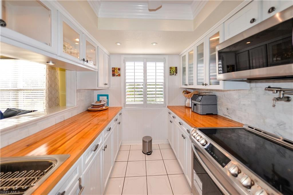 Galley kitchen featuring white cabinetry, stainless steel appliances, wood-finish countertops, tiled flooring, and a window with plantation shutters