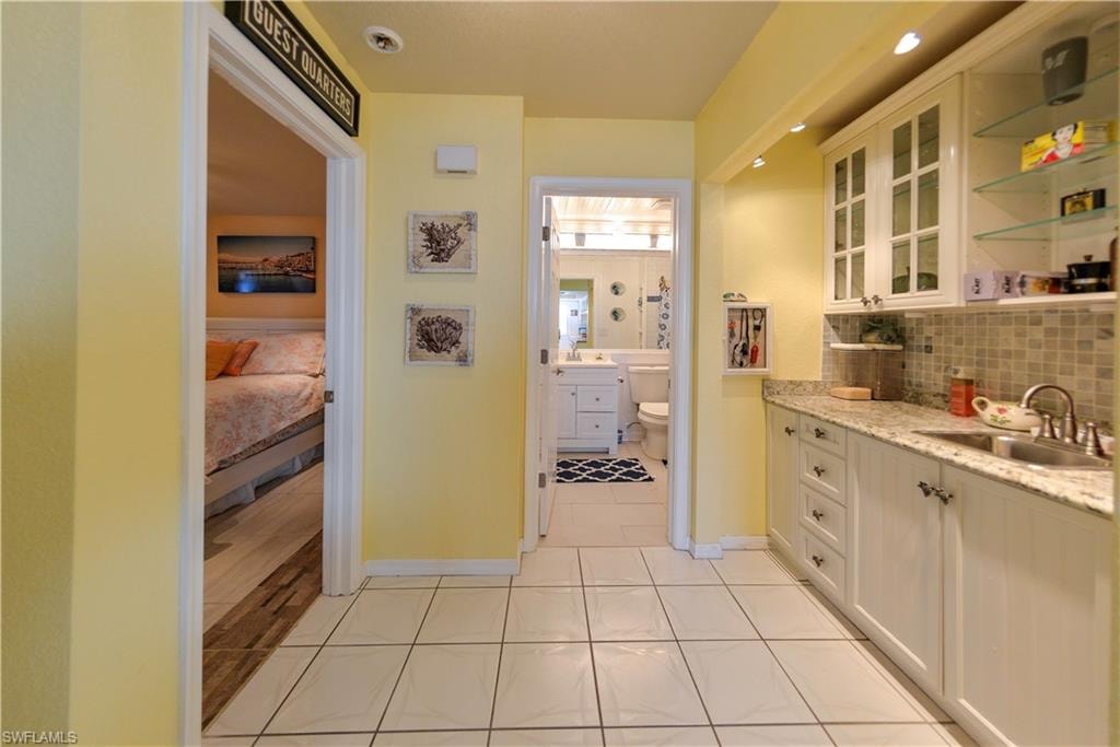 Hallway featuring white square tile flooring, a built-in wet bar with granite countertops, white cabinetry, a stainless steel sink, and glass shelving