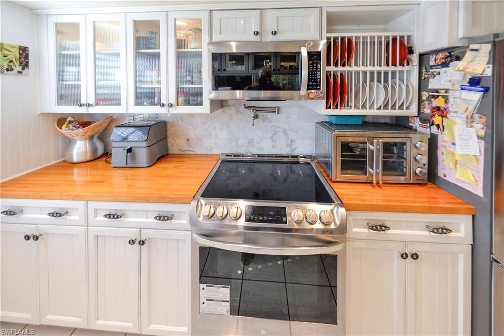 Modern kitchen featuring a stainless steel range with a flat cooktop, white shaker-style cabinetry, butcher block countertops, a tile backsplash, and upper cabinets with glass-paneled doors