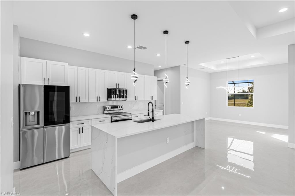 Kitchen with decorative light fixtures, appliances with stainless steel finishes, light stone counters, white cabinets, and an island with sink