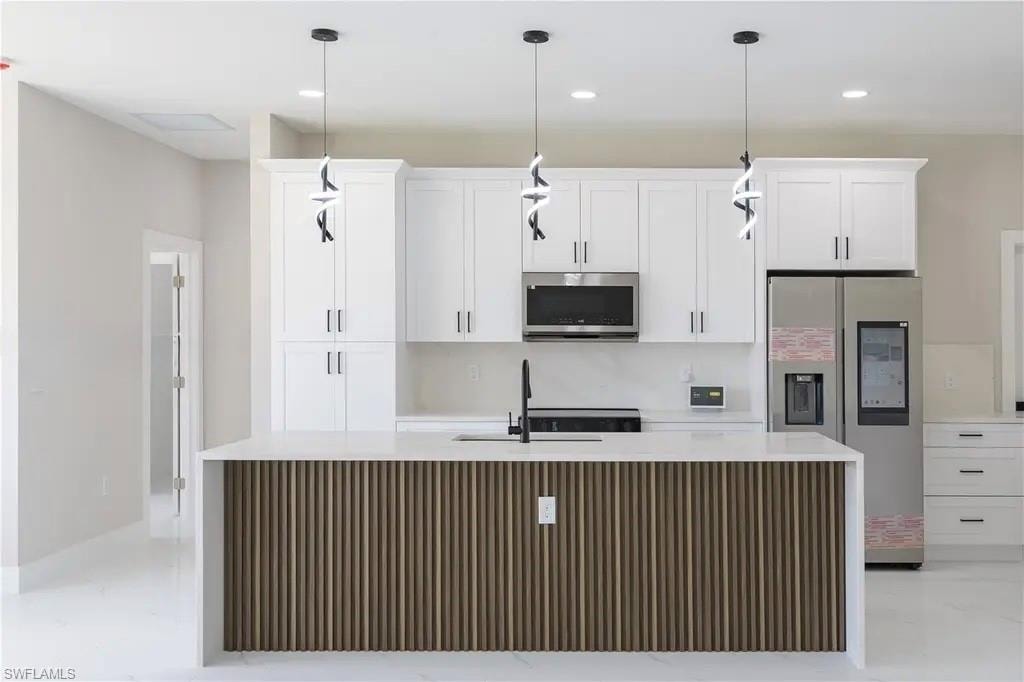 Kitchen with stainless steel appliances, an island with sink, white cabinetry, light stone counters, and decorative light fixtures