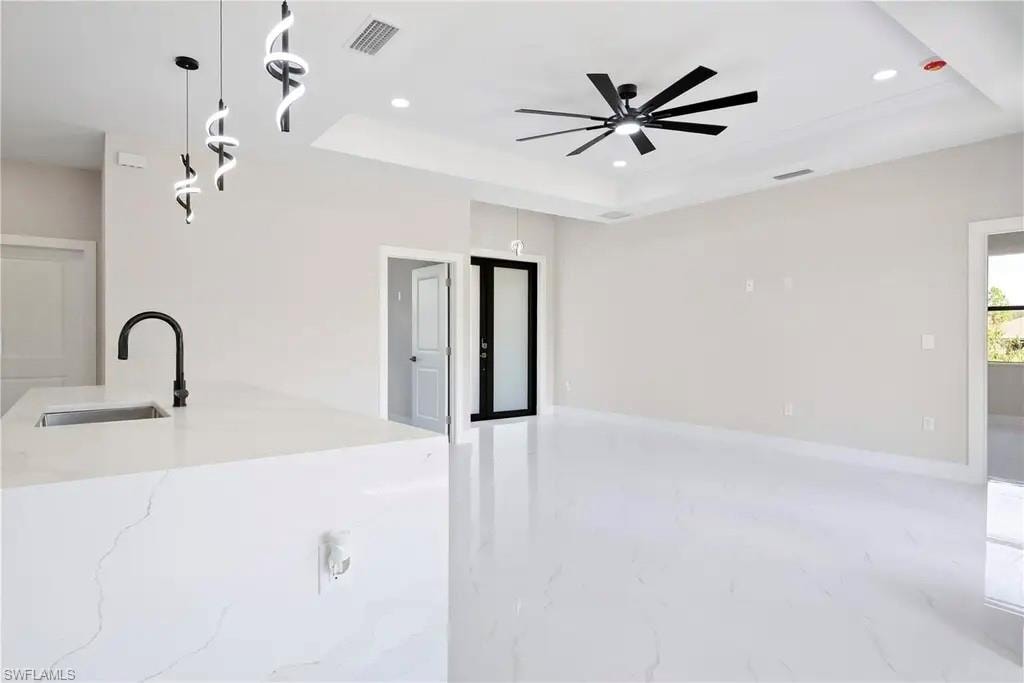 Kitchen with hanging light fixtures, a tray ceiling, light stone counters, ceiling fan, and an island with sink