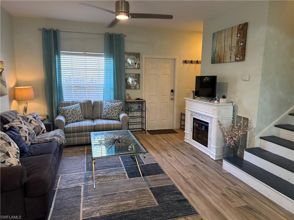 Wood-finish flooring, ceiling fan with integrated lighting, window with blinds, white entry door, and a decorative electric fireplace