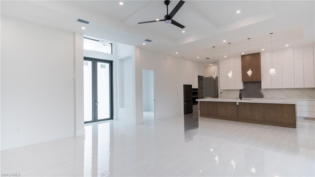 Spacious open-concept living area featuring reflective white tile flooring, a modern kitchen island with pendant lighting, black ceiling fan, recessed lighting, and double-door entry