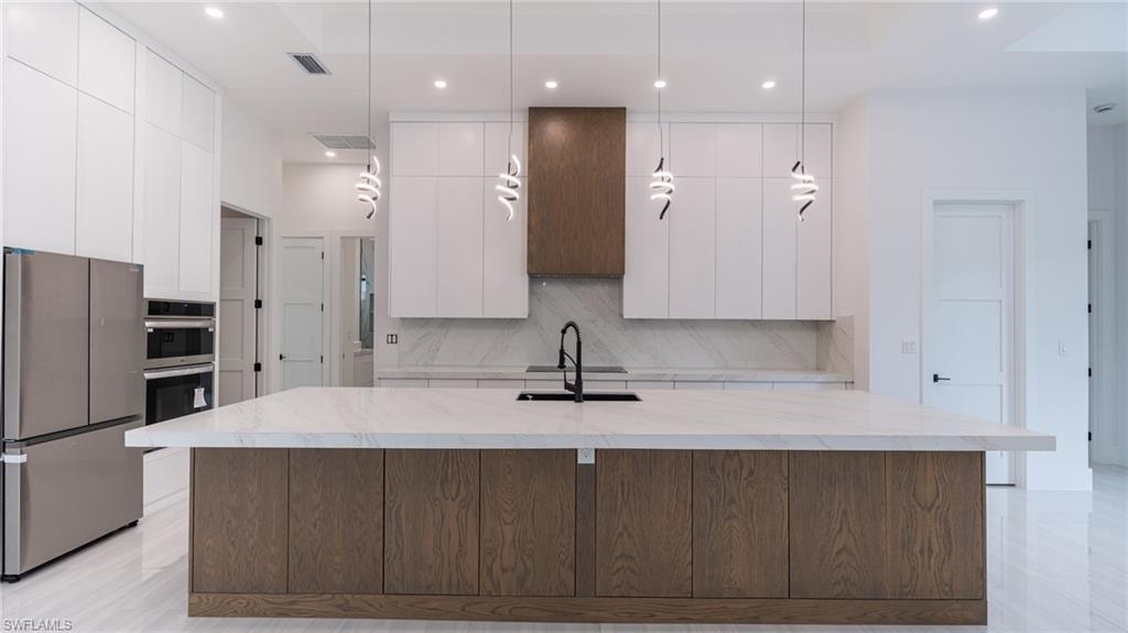 Gourmet kitchen featuring a large central island with wood-finish cabinetry, stainless steel appliances, white flat-panel upper cabinetry, a coordinating wood-finish range hood, and pendant lighting fixtures