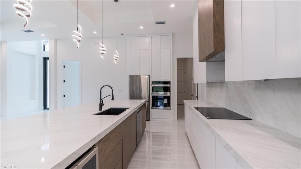 Spacious kitchen featuring a large central island with an undermount sink, quartz countertops, integrated stainless steel appliances, and sleek white cabinetry with wood-finish accents