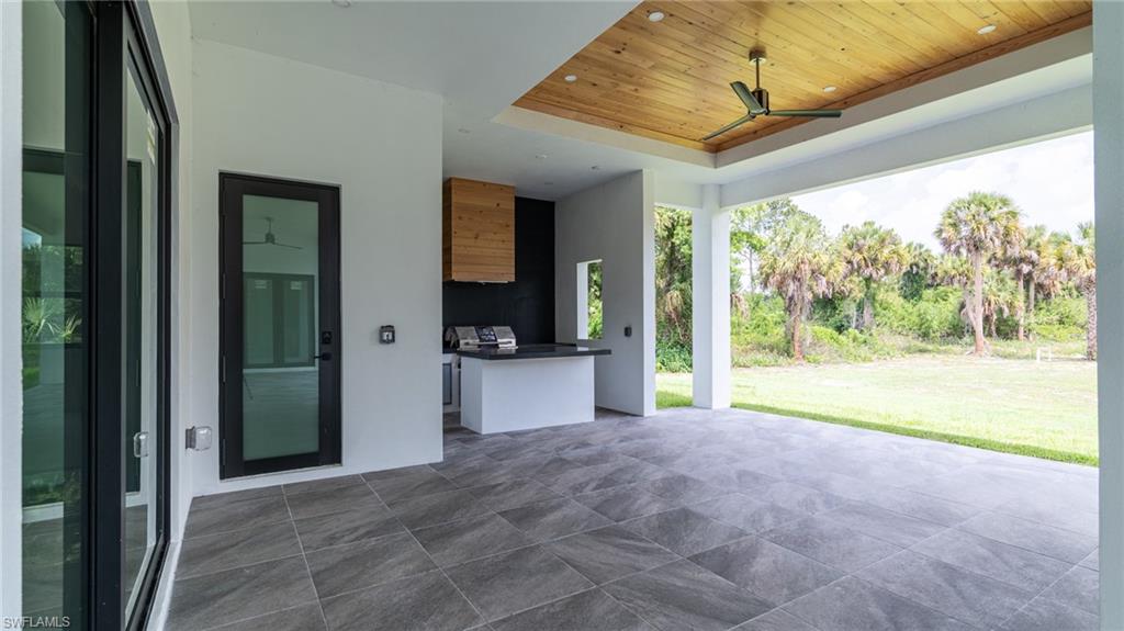 Expansive covered patio featuring large format tile flooring, a wood-paneled ceiling with a ceiling fan, and an integrated outdoor kitchen with a stone countertop