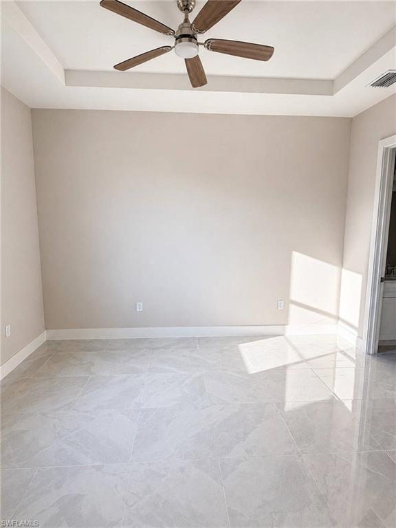 Spacious room featuring a tray ceiling with a ceiling fan, light gray wall paint, white baseboards, and large format light grey tile flooring