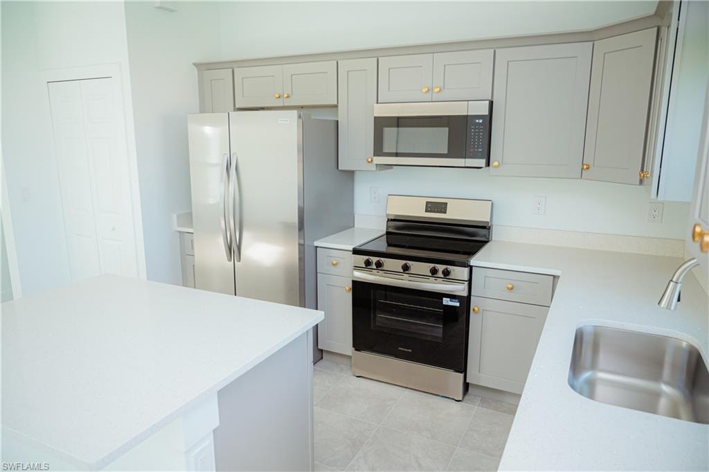 Modern kitchen featuring light gray cabinetry with gold-toned hardware, white solid-surface countertops, and a stainless steel appliance suite including a refrigerator, range, and microwave