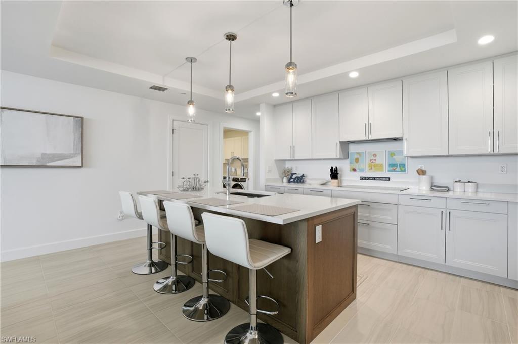 Kitchen featuring a raised ceiling, an island with sink, a kitchen breakfast bar, white cabinetry, and decorative light fixtures - Virtually Edited Image