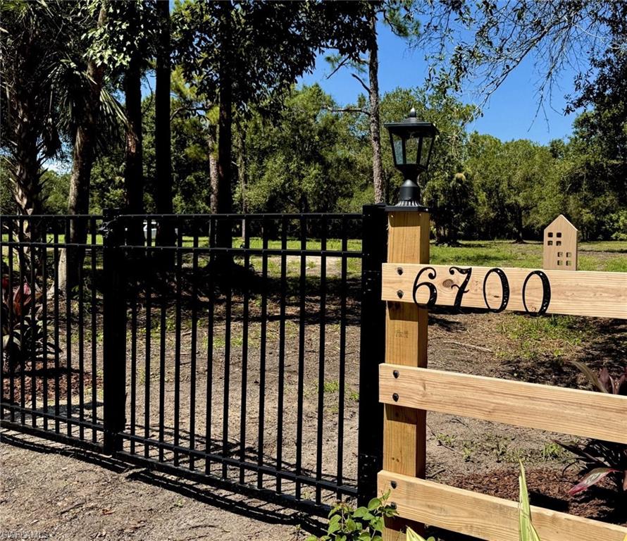 Welcoming entrance featuring a black metal driveway gate with decorative finials, natural wood-finish address marker displaying "6700," and mature tree-lined property with expansive grounds and wooden ranch-style fencing
