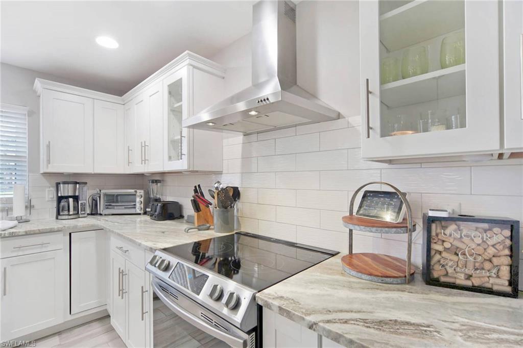 Kitchen featuring glass insert cabinets, stainless steel electric range oven, wall chimney range hood, light stone countertops, and white cabinetry